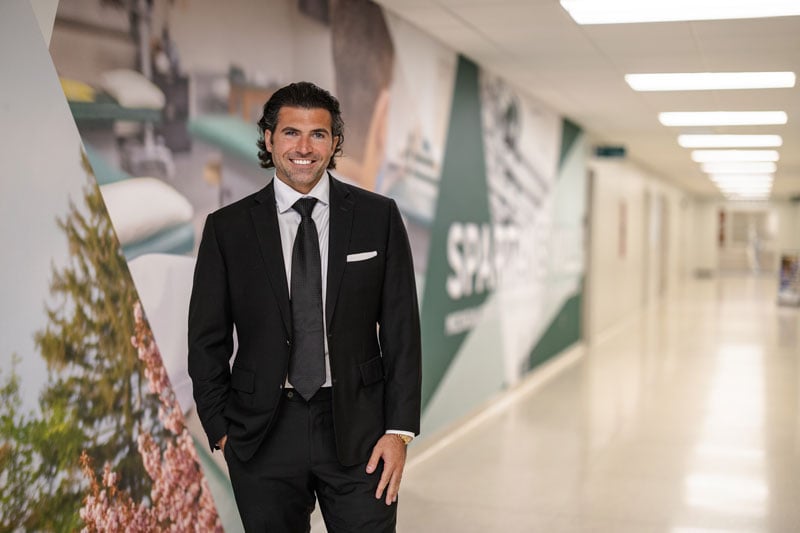 A person smiles while standing in a brightly lit hallway with modern decor and wall graphics. They are wearing a black suit with a white shirt, black tie, and a pocket square. The hallway features large, colorful murals, including images of trees and medical equipment, with part of the word "Spartans" visible on the wall. The space is clean and contemporary, with a long corridor extending into the background.