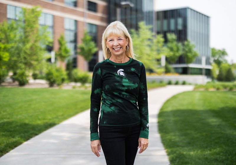 A person smiles while standing outdoors on a paved path lined with green grass and trees. They are wearing a dark green long-sleeve shirt featuring the Michigan State University Spartan helmet logo and black pants. Behind them, modern brick and glass buildings are visible, set against a bright, sunny sky.