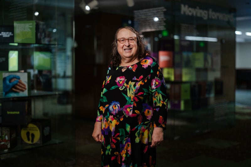 A person stands smiling in a well-lit indoor space with a contemporary exhibit visible in the background. They are wearing glasses and a long, black dress adorned with colorful floral patterns. The display behind them features text panels, illuminated signage, and exhibit cases with various objects and graphics. The word "Moving forward" is partially visible in the background.