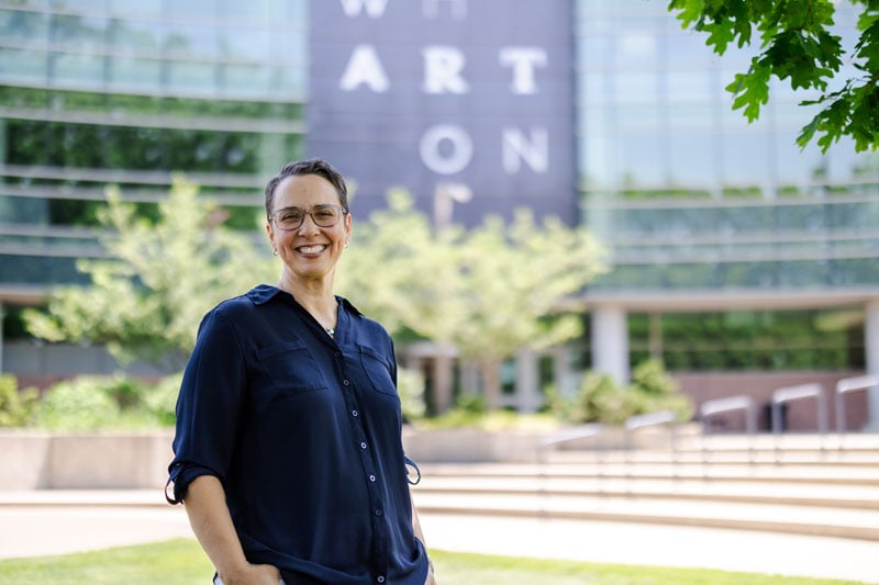 A person stands smiling outdoors in front of the Wharton Center for Performing Arts. They are wearing glasses and a navy blue button-up shirt, with their hands in their pockets. The background features the building's glass facade, trees, and a portion of a large sign with the word "Wharton" visible. The scene is bright and sunny, with a green leafy tree branch framing the top corner.