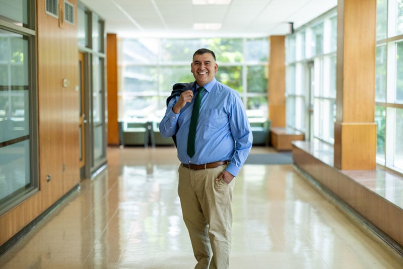 A person smiles while standing in a sunlit hallway with large windows. They are dressed in a light blue button-up shirt, a green tie, and khaki pants, holding a dark jacket over their shoulder. The hallway features wooden paneling, polished floors, and views of greenery outside through the windows, creating a bright and inviting atmosphere.
