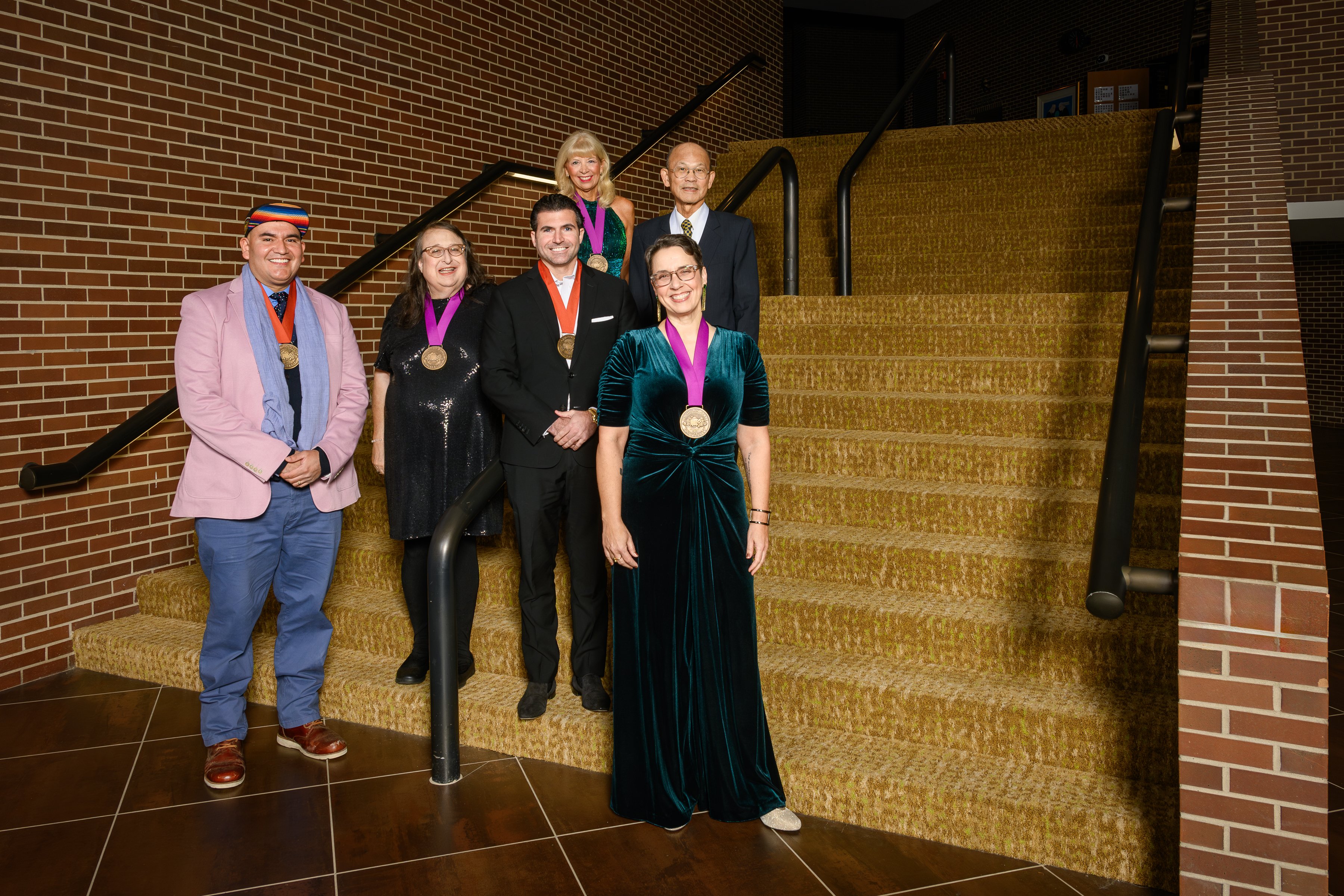  Six people wearing medals pose on a carpeted staircase in formal and semi-formal attire. The brick walls and dim lighting create an elegant setting.