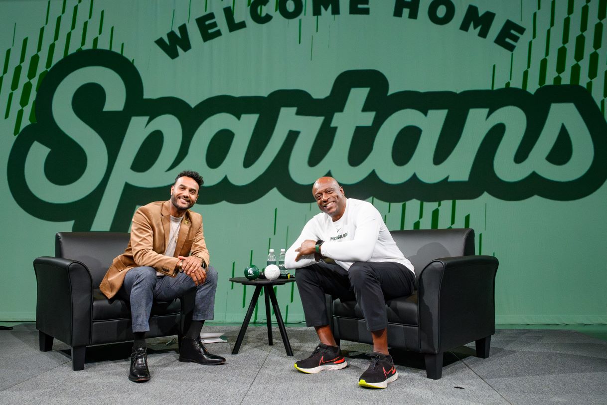 Two men sit on a stage with a large green banner that reads "Welcome Home Spartans" in the background. One is dressed in a tan blazer, and the other in a white Michigan State shirt, both smiling at the camera.