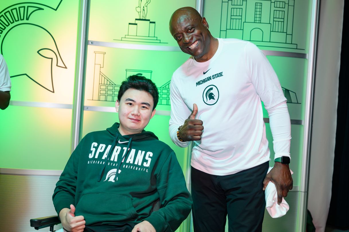 Two individuals pose for a photo at a Michigan State University event. One is seated, wearing a green Spartans hoodie, and the other stands beside them, smiling and giving a thumbs-up. The backdrop features Spartan-themed graphics and green lighting.