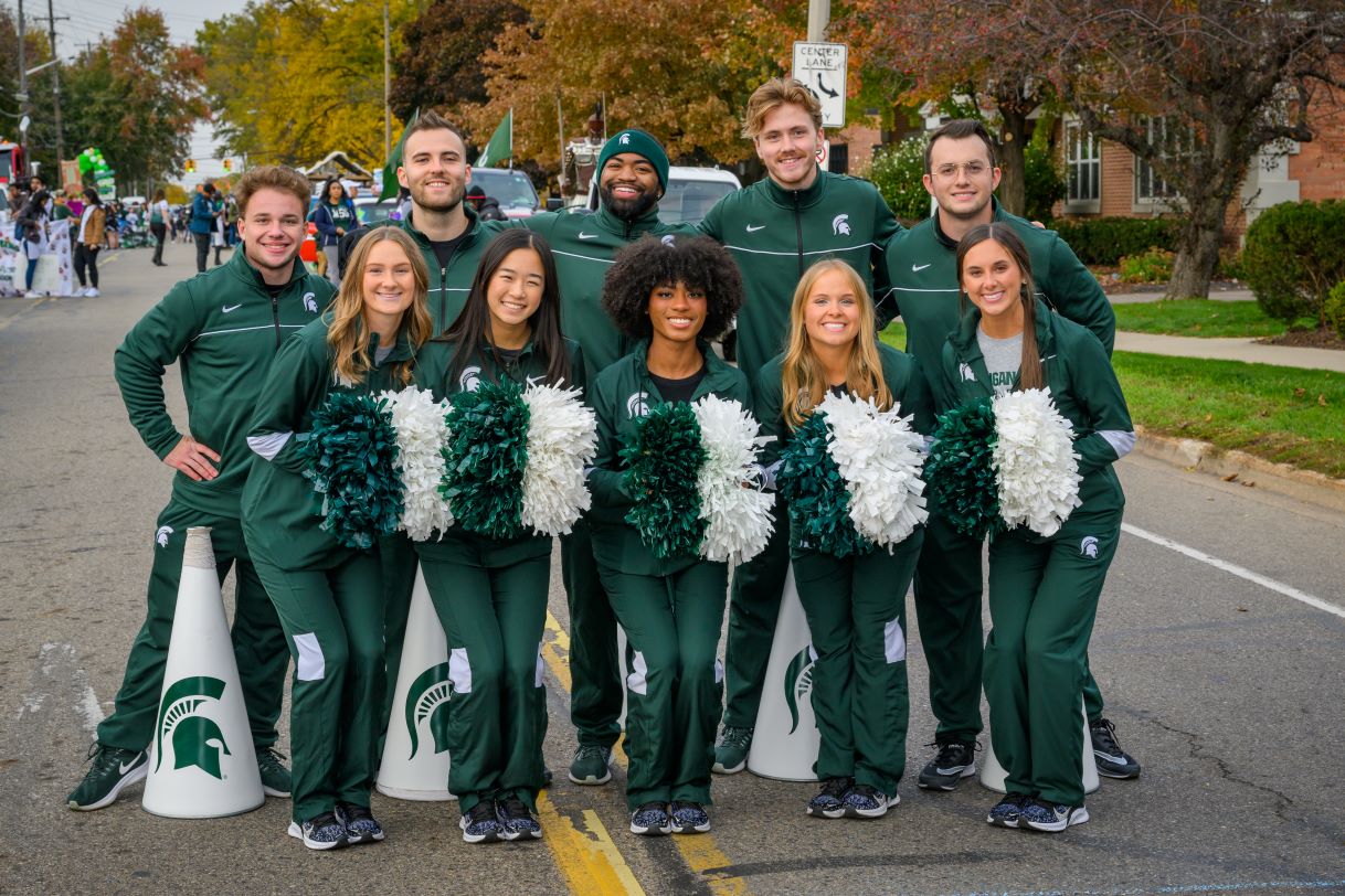 Michigan State University cheerleaders pose in a group on a parade route. They are dressed in matching green outfits, holding green and white pom-poms, with Spartan megaphones placed in front of them. The fall scenery adds warmth to the background.