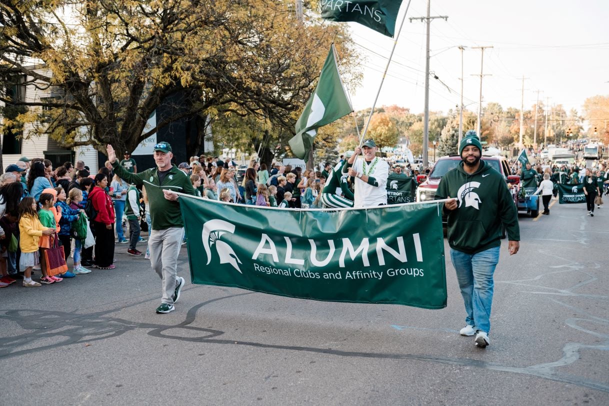A group of Michigan State University alumni carry a large green banner that reads "Alumni Regional Clubs and Affinity Groups" during a parade. They are surrounded by a crowd of spectators along a tree-lined street with a festive autumn atmosphere.