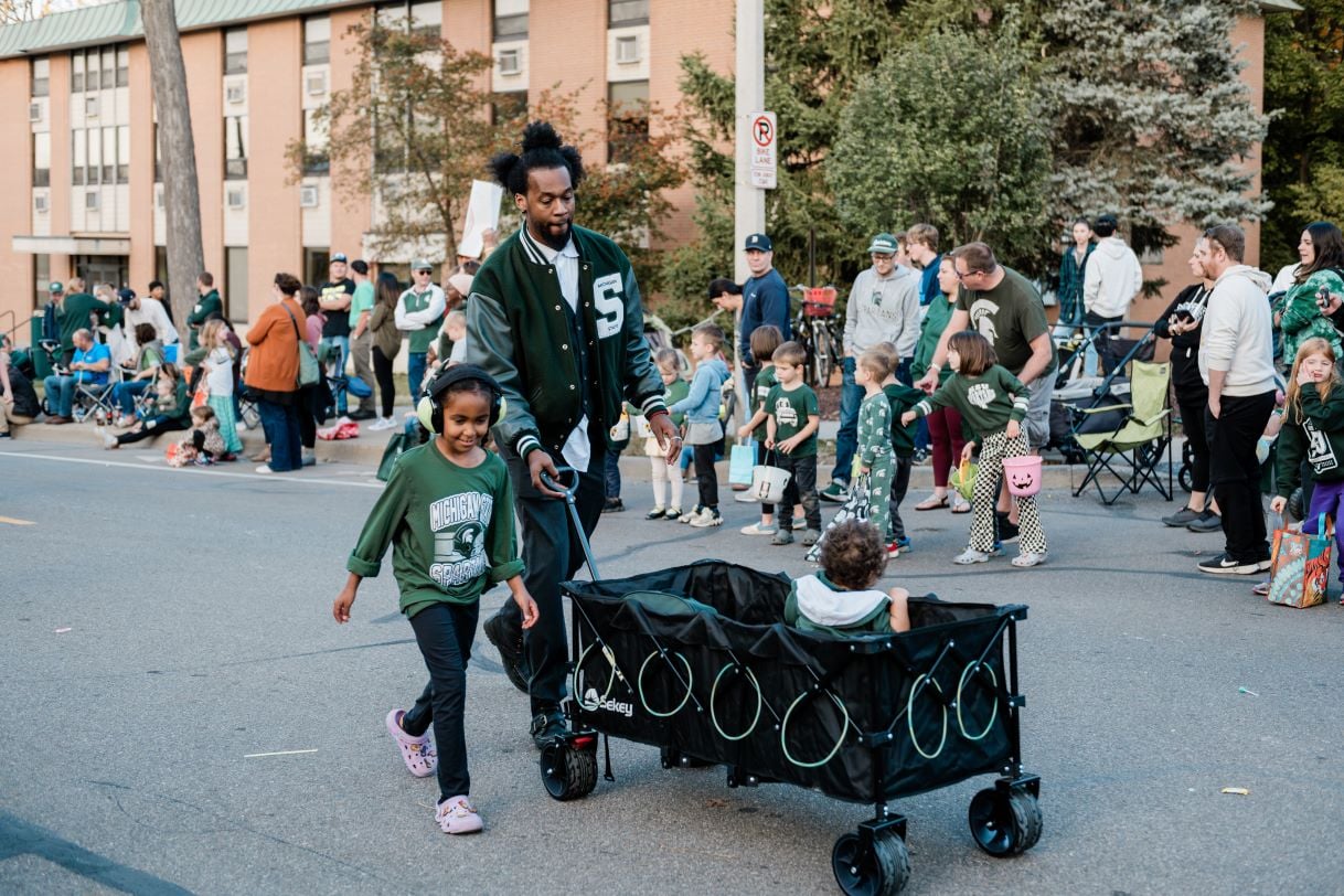 A Michigan State University parade attendee wearing a green varsity jacket walks alongside a young girl in a Spartan shirt and headphones. The child pulls a wagon with another young child seated inside. Spectators line the street, enjoying the festive atmosphere.