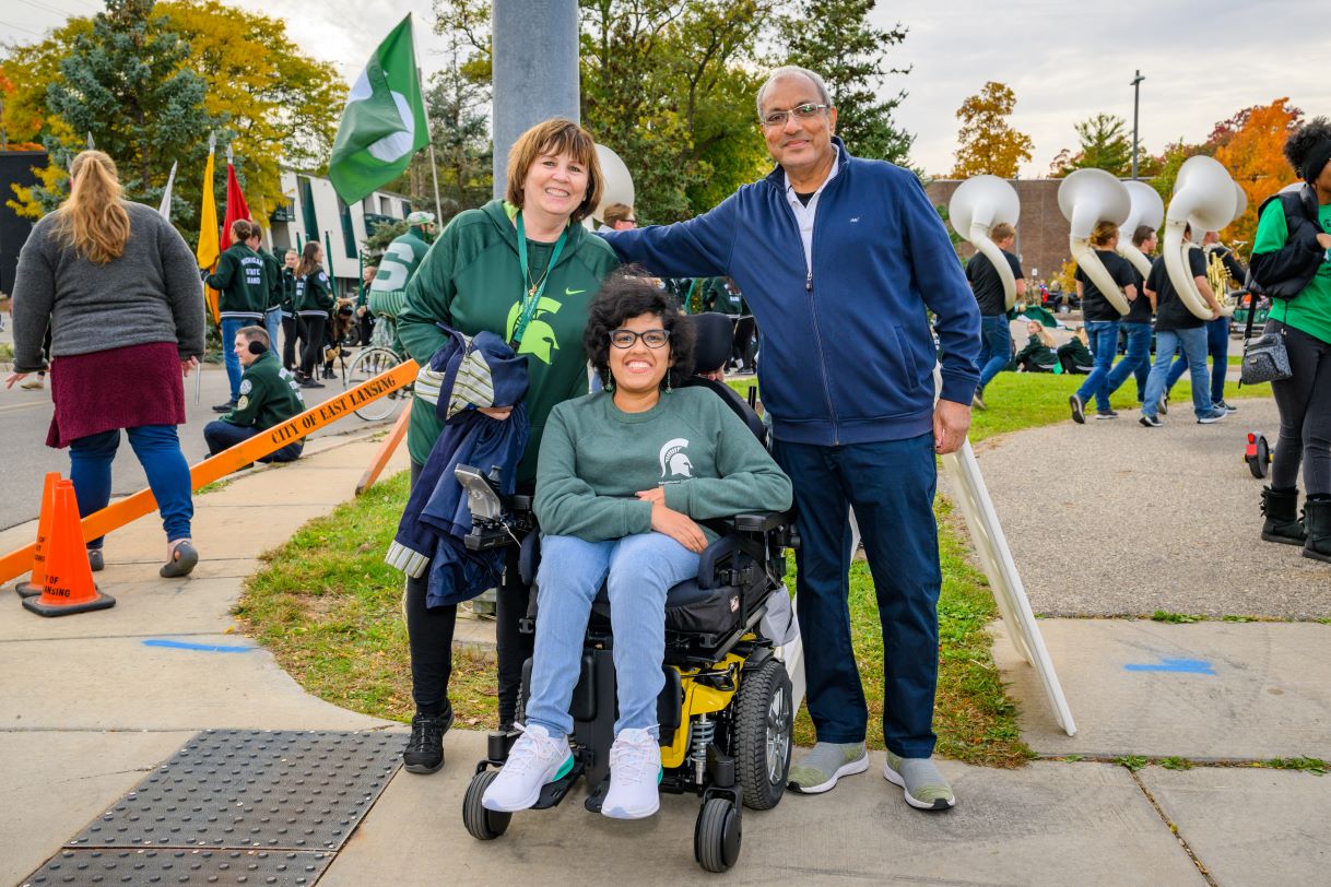Three smiling individuals pose together at the Michigan State University Homecoming Parade. One person is seated in a wheelchair wearing a Spartan sweatshirt, and two others stand beside them in Spartan-themed clothing with a band performing in the background.