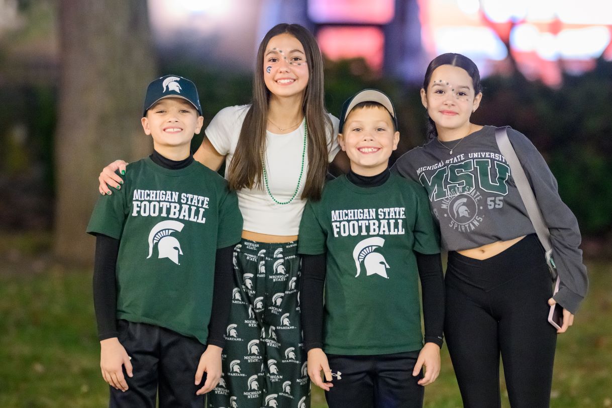 A group of four young attendees poses together, smiling and dressed in Michigan State University-themed apparel, including "MSU" and "Michigan State Football" shirts. The backdrop is a dimly lit evening setting with vibrant lights.