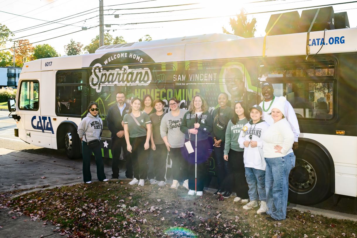 A group of Michigan State University alumni and students stands in front of a decorated CATA bus with the words "Welcome Home Spartans" and "Homecoming" on the side. The sunny day and autumn leaves create a vibrant backdrop.