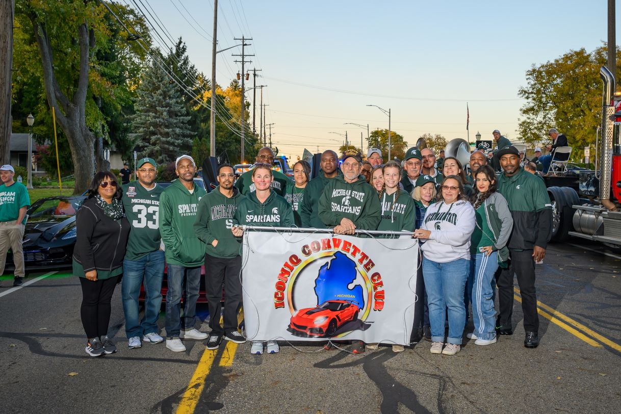A group of Michigan State University alumni and community members stand together holding a banner for the "Iconic Corvette Club" during the parade. Many wear Spartan-themed clothing, and classic cars are visible in the background.