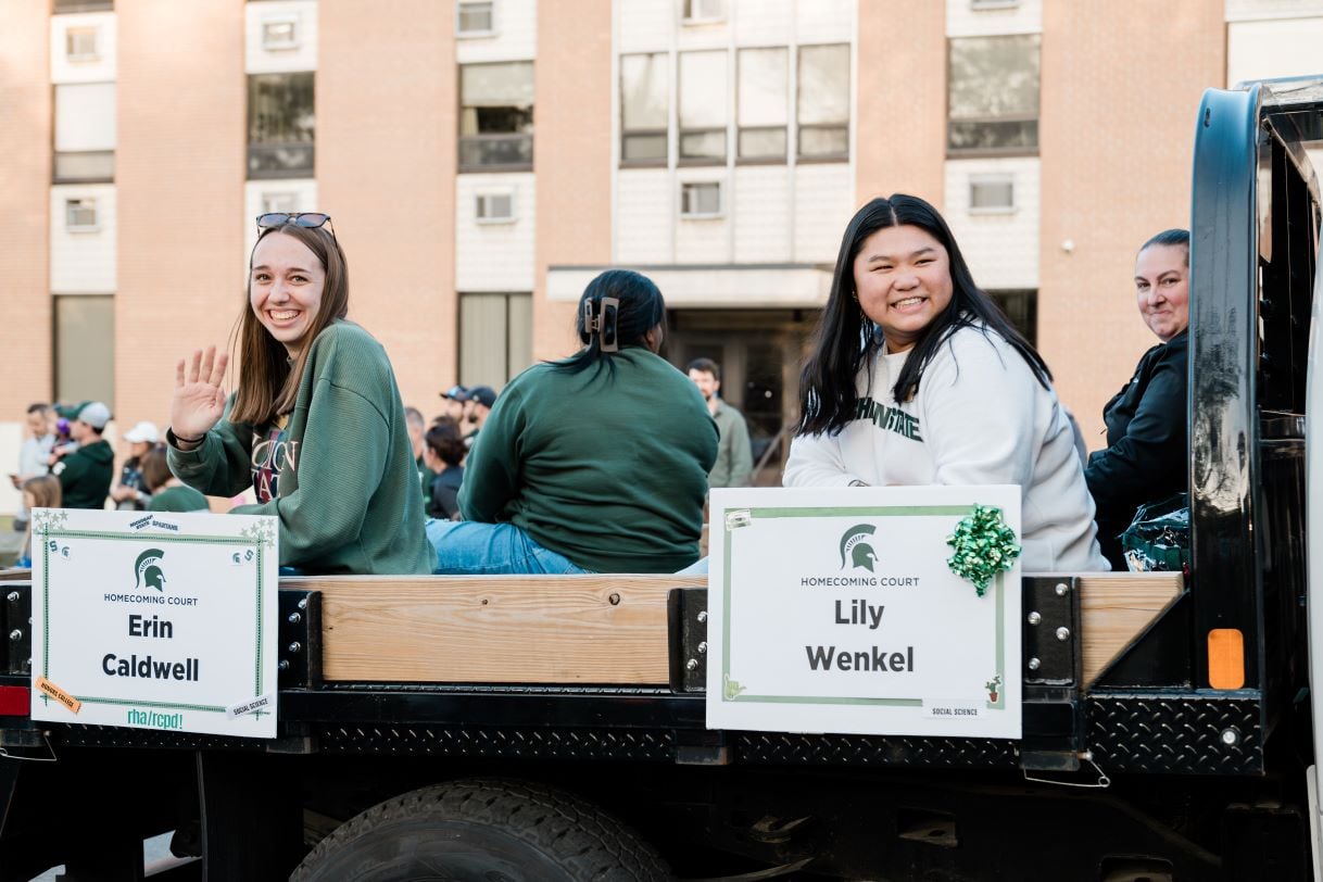 Two smiling individuals, Erin Caldwell and Lily Wenkel, sit on a parade float labeled "Homecoming Court." Both are dressed in green and white Michigan State University apparel and wave to the crowd during the festive parade.