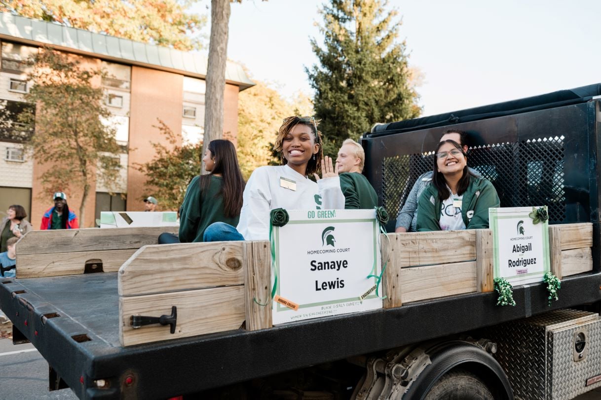 Sanaye Lewis and Abigail Rodriguez, members of the Michigan State University Homecoming Court, wave from a float decorated with green and white signage during the parade. Both are dressed in Spartan apparel and smiling brightly.