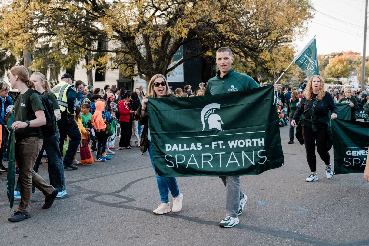 Members of the Dallas-Ft. Worth Spartans alumni group hold a large green banner during the Michigan State University Homecoming Parade. The festive scene includes a crowd of spectators and autumn trees.
