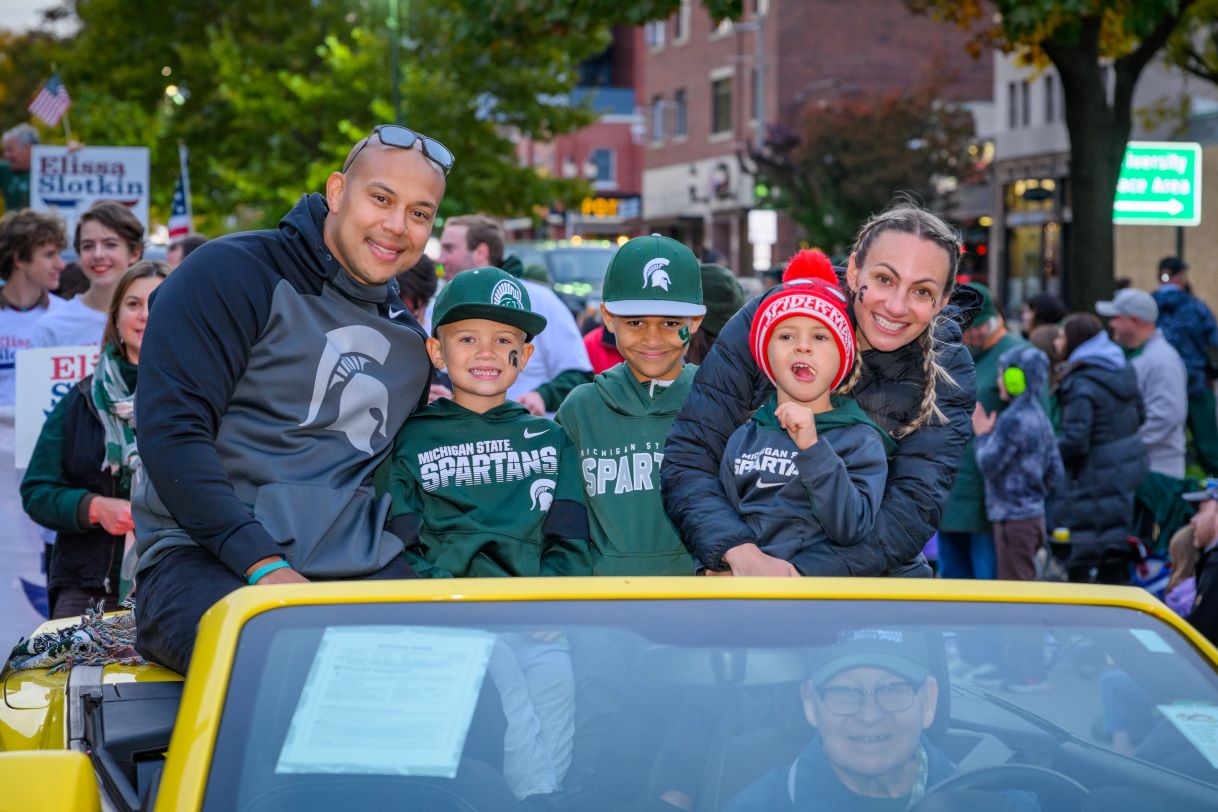 A family of five smiles from a bright yellow car participating in the Michigan State University Homecoming Parade. The family wears Spartan-themed clothing, including green hoodies and caps, while the parade crowd watches from the sidewalk.