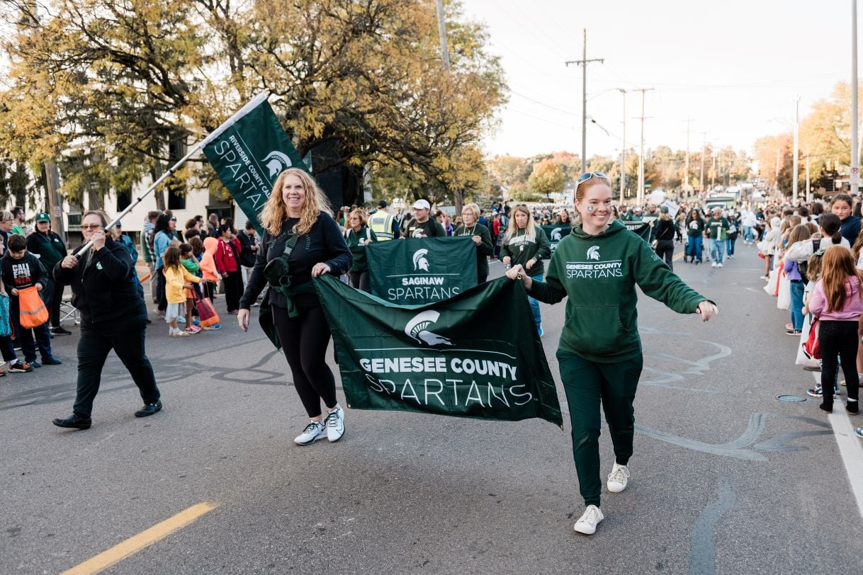 Members of the Genesee County Spartans alumni group carry a large green banner during the Michigan State University Homecoming Parade. Spectators line the street, and trees with fall foliage frame the scene.