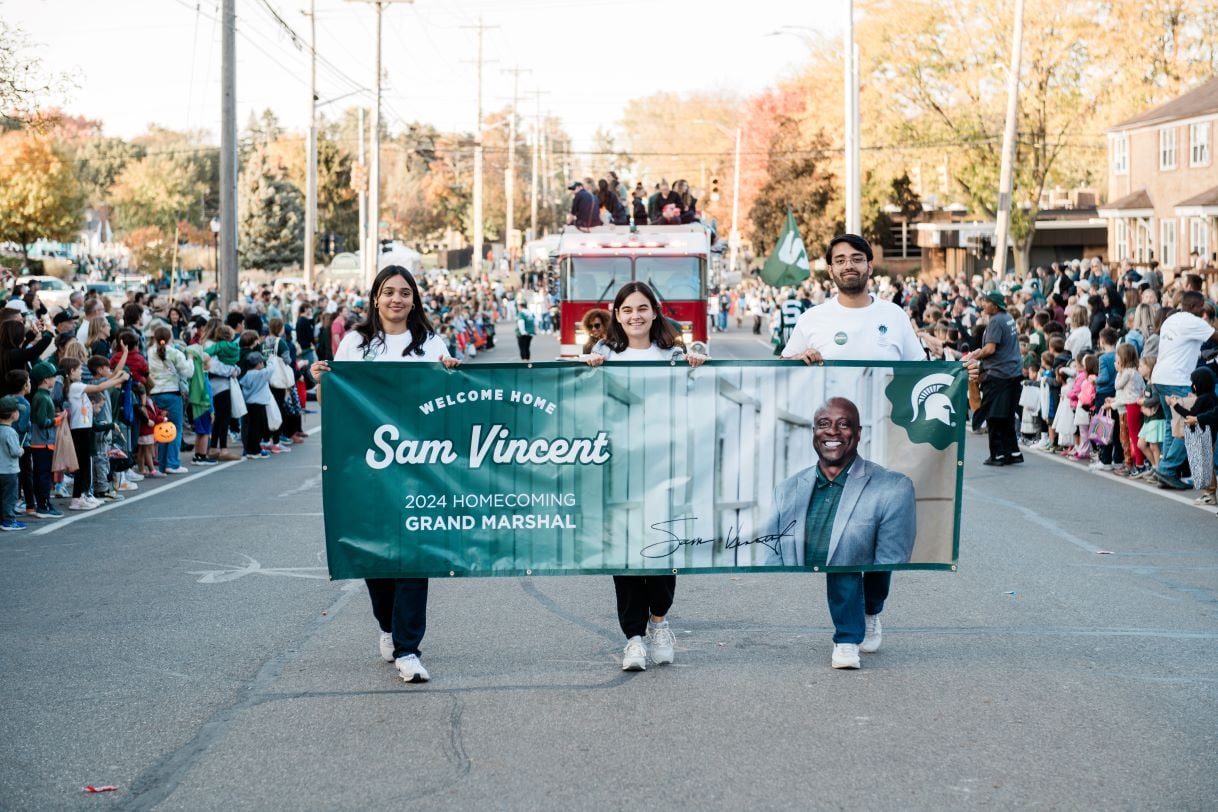 Four individuals carry a large green banner reading "Welcome Home Sam Vincent, 2024 Homecoming Grand Marshal" during the Michigan State University Homecoming Parade. A firetruck and spectators fill the background.