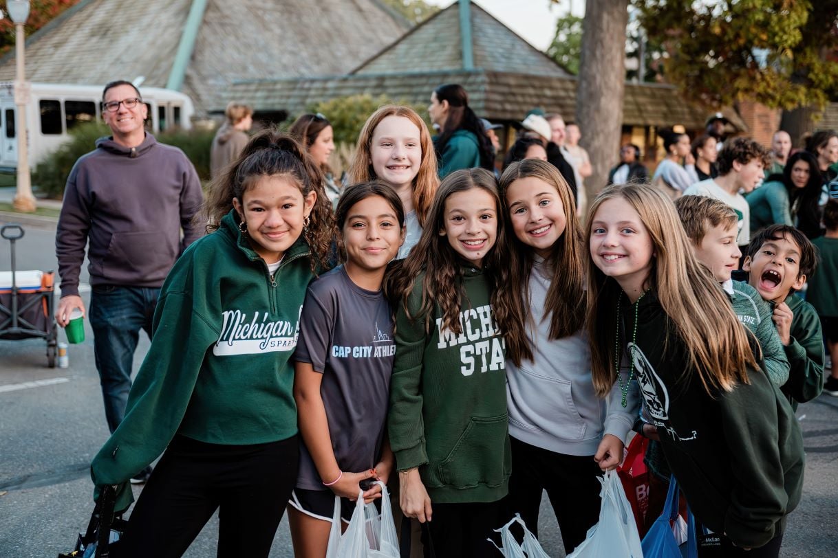 A group of smiling kids pose together at the Michigan State University Homecoming Parade. Many are wearing Spartan-themed clothing, including hoodies and shirts, and hold candy bags. The festive crowd and trees are visible in the background.