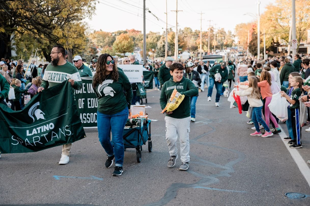 Members of the Latino Spartans group walk in the Michigan State University Homecoming Parade, carrying a green banner with the Spartan logo. A young boy smiles as he hands out candy to the cheering crowd lining the street.