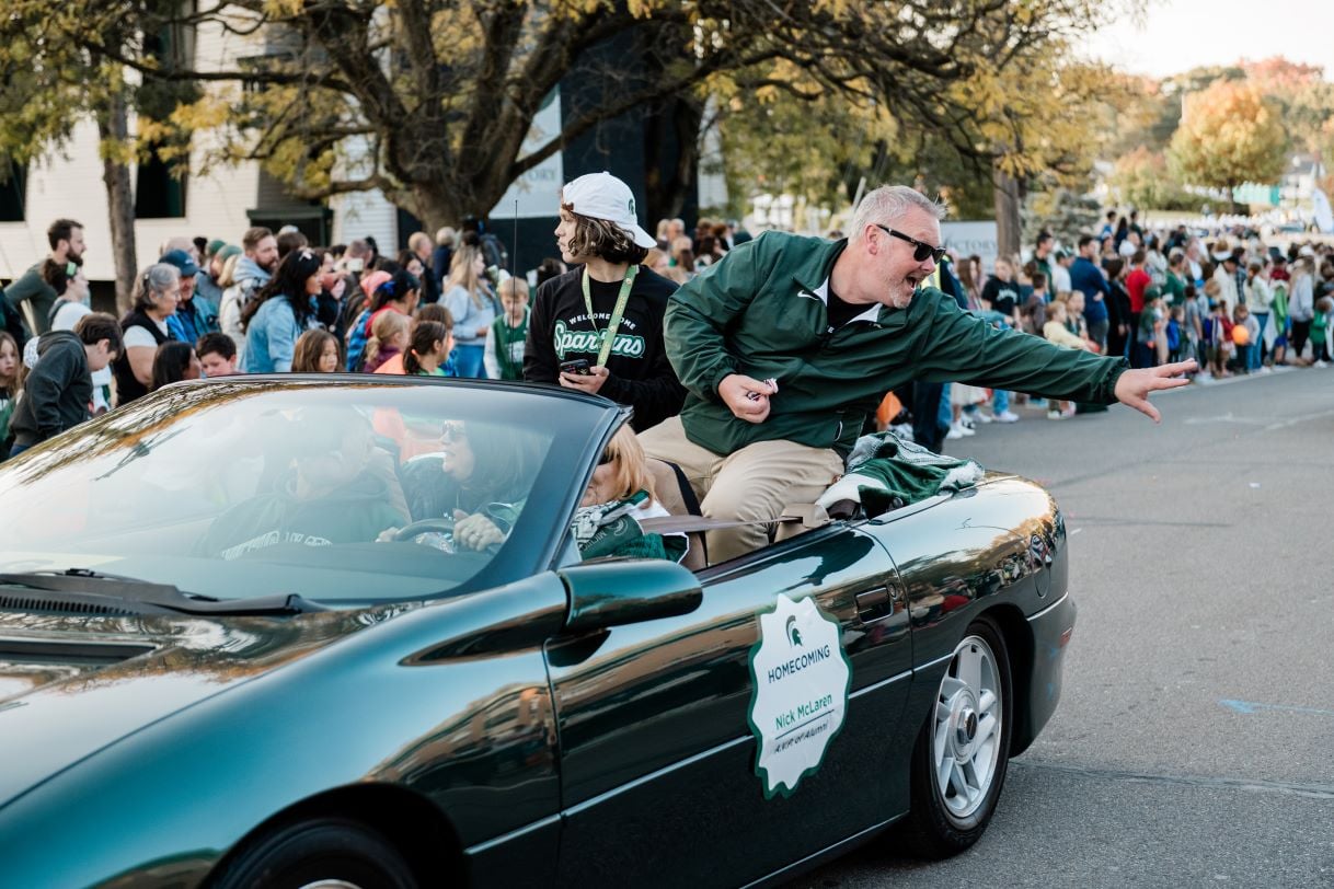 Nick McLaren waves and interacts with the crowd from a green convertible in the Michigan State University Homecoming Parade. The car displays a sign with his name and "Homecoming" on the side, while spectators watch from the sidewalk.