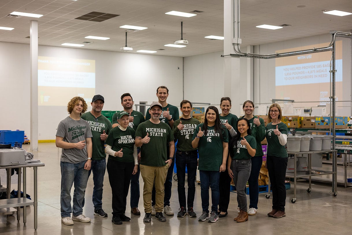 A group of Austin Spartans in Michigan State shirts giving thumbs up inside a food bank, standing in front of donation stations.