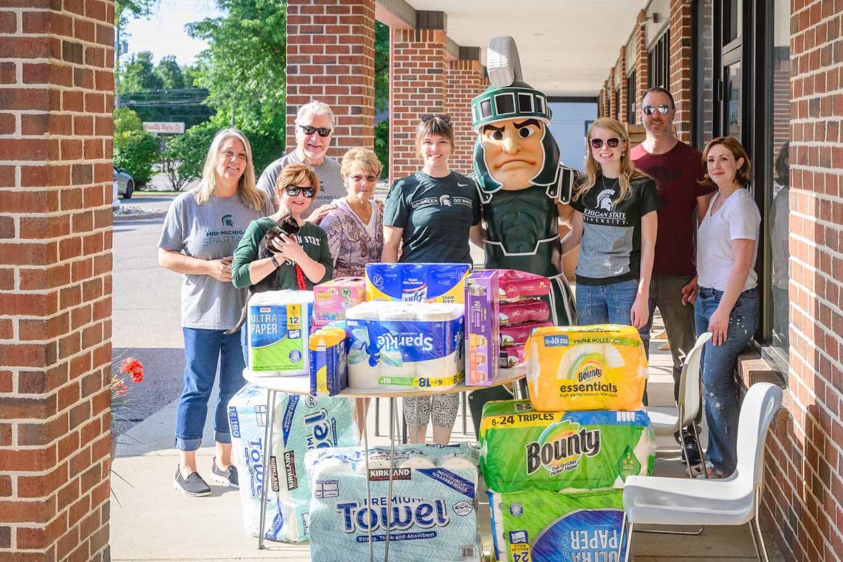 Mid-Michigan Spartans, including Sparty mascot, pose outside with donations of paper products and pet supplies for a cat adoption center.