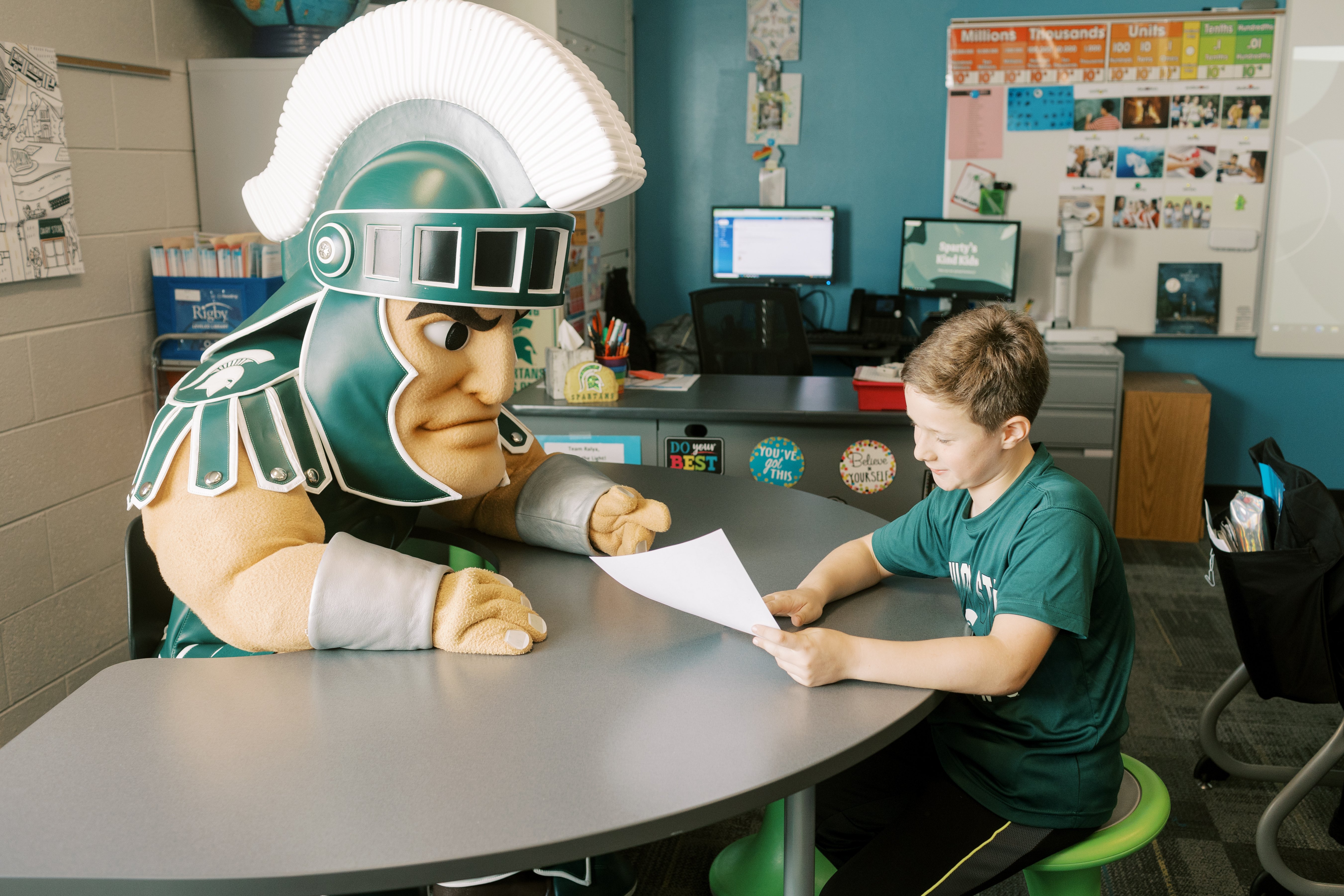  A child sits at a classroom table, reading a paper while MSU's Sparty mascot leans in to listen. The room features a desk, computer, and colorful decorations.