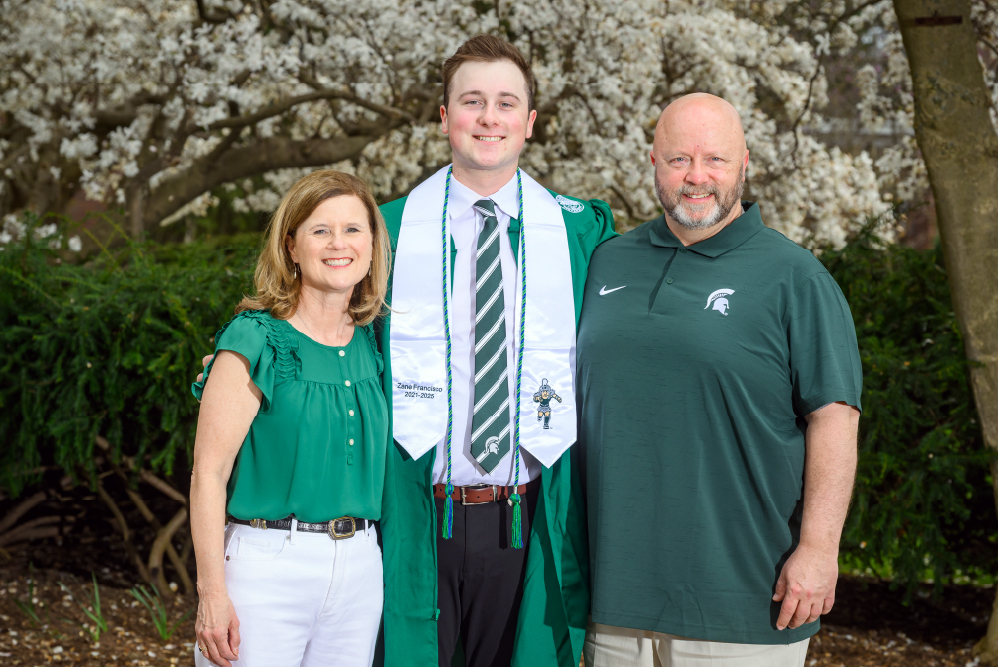 Graduate Zane Francisco stands proudly between his parents, all smiling in front of blooming trees. Zane is dressed in his green cap and gown with a white stole, while his parents wear Spartan green in celebration.