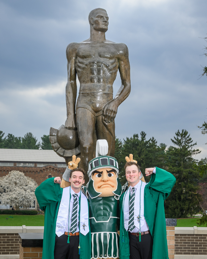 Graduates Zach O’Brien and Zane Francisco pose with the Sparty mascot in front of The Spartan statue at Michigan State University. All three strike celebratory poses, with the iconic bronze figure towering behind them.