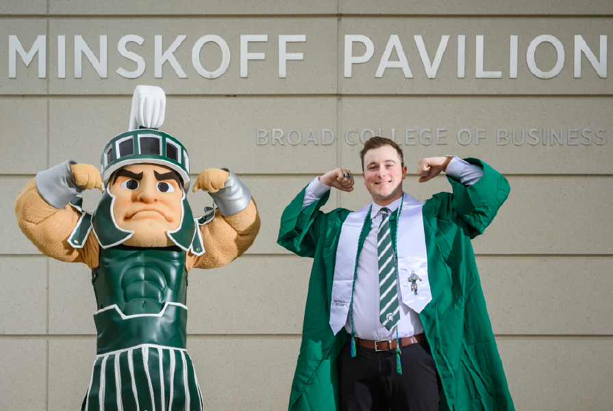 Zane Francisco, in graduation attire, poses proudly with the Sparty mascot in front of Minskoff Pavilion at the Broad College of Business, both flexing their arms in a show of Spartan pride.