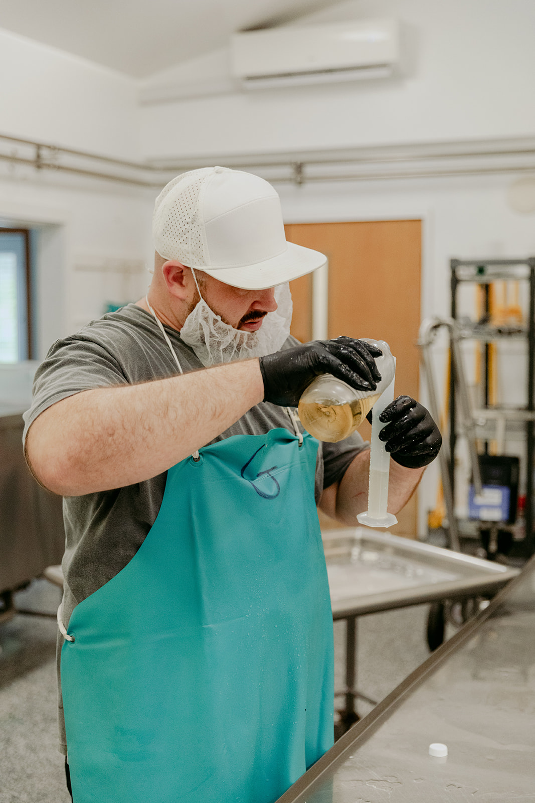 Gary Smith in a production facility, wearing a teal apron and white cap, carefully measuring liquid for cheese making.