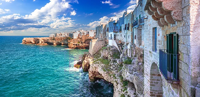 A breathtaking coastal view of Polignano a Mare in Italy, showcasing historic stone buildings perched on rugged cliffs overlooking the vivid turquoise waters of the Adriatic Sea. The sunlight illuminates the scene under a partly cloudy sky, highlighting the dramatic contrast between the natural cliffs and the architecture. The waves crash against the rocks, creating a sense of movement and energy.