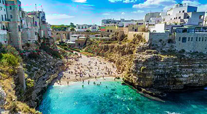 A stunning coastal view of Polignano a Mare in southern Italy, featuring a small sandy beach surrounded by rugged cliffs and turquoise waters. The historic town's whitewashed buildings sit perched atop the cliffs, offering a picturesque backdrop under a bright blue sky with scattered clouds. Visitors enjoy the beach and swim in the clear waters, capturing the charm and beauty of this Mediterranean destination.
