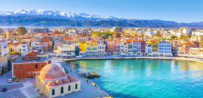 A picturesque view of the Venetian harbor in Chania, Crete, featuring colorful buildings lining the waterfront. The turquoise water reflects the vibrant facades, while the historic mosque of Kioutsouk Hassan is visible in the foreground. Snow-capped mountains rise majestically in the background under a clear blue sky, adding to the stunning coastal scenery.