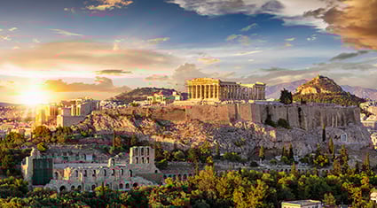 A stunning view of the Acropolis in Athens, Greece, at sunset. The Parthenon sits atop the ancient hill, surrounded by historic ruins and lush greenery. The cityscape of Athens spreads out in the background, bathed in the warm golden light of the setting sun, with a dramatic sky filled with clouds adding depth to the scene.