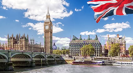 A vibrant daytime view of London, England, featuring Big Ben and the Houses of Parliament along the River Thames. Westminster Bridge crosses the river, with boats and a British Union Jack flag waving prominently in the foreground. The blue sky is dotted with fluffy white clouds, creating a lively and iconic scene of the city.