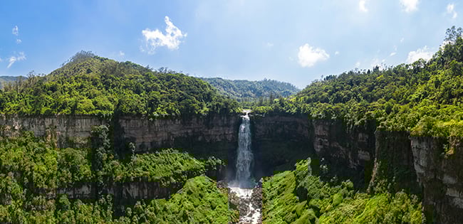 A stunning view of a tall waterfall cascading into a lush green valley surrounded by cliffs and dense forest, with a bright blue sky and scattered white clouds above.