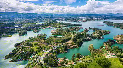 A breathtaking aerial view of a lush landscape featuring interconnected lakes, green hills, and scattered houses. The winding roads and vibrant greenery create a serene and picturesque scene, with a partly cloudy sky adding depth to the backdrop. The vibrant colors of nature dominate the image, showcasing the beauty of the region.