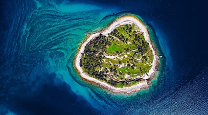 An aerial view of a small, heart-shaped island surrounded by the vibrant turquoise waters of the Adriatic Sea in Croatia. The island is covered with lush greenery and edged by a rocky shoreline, creating a striking contrast with the deep blue of the surrounding water. The scene showcases the unique natural beauty of Croatia's coastal region.