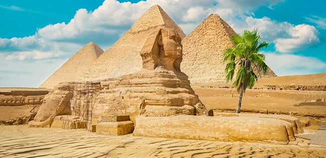 The Great Sphinx of Giza with the iconic Pyramids of Giza in the background, located in Egypt. The ancient limestone statue of the Sphinx, featuring a lion's body and a human head, stands prominently on a sandy plateau. Behind it, the three grand pyramids rise majestically under a bright blue sky with scattered white clouds. A lone palm tree adds a touch of greenery to the arid desert landscape.