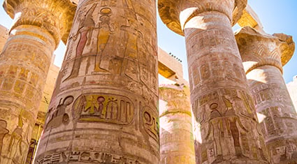 Ancient stone columns at the Karnak Temple Complex in Luxor, Egypt, adorned with intricate hieroglyphics and colorful carvings depicting Egyptian deities and symbols. The towering columns are illuminated by sunlight, showcasing their detailed artwork and faded hues. The open sky is visible through gaps in the structure, adding to the historical and majestic ambiance.