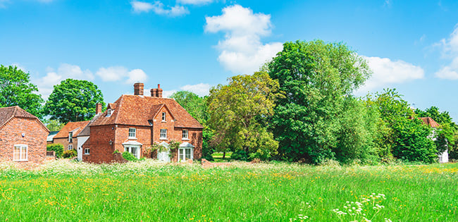 A charming countryside scene in England featuring a red-brick house with a tiled roof surrounded by lush greenery and colorful trees. The house is set amidst a vibrant meadow filled with wildflowers under a bright blue sky with fluffy white clouds, creating a serene and idyllic rural setting.