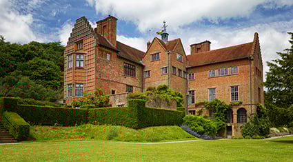 A historic brick manor house in England surrounded by lush green gardens and manicured hedges. The building features a red-tiled roof, chimneys, and ornate architectural details. The vibrant lawn in the foreground and the wooded backdrop create a serene and picturesque setting under a partly cloudy blue sky.
