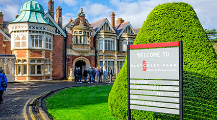 The entrance to Bletchley Park in England, featuring a historic red-brick mansion with ornate architecture, including pointed gables and a distinctive green domed turret. A sign in the foreground reads "Welcome to Bletchley Park - National Code Centre," set against a neatly trimmed hedge and green lawn. A group of visitors is gathered near the entrance under a sunny blue sky.