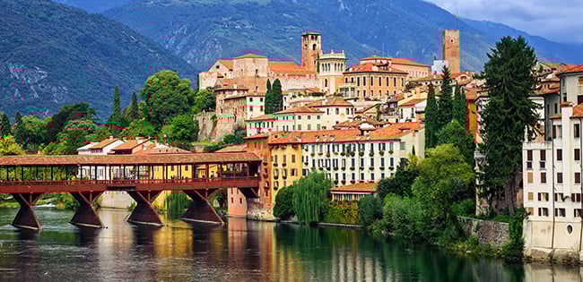 A charming view of Bassano del Grappa in northern Italy, featuring colorful historic buildings lining the Brenta River. The iconic wooden Ponte degli Alpini bridge spans the river, with lush greenery and rolling hills in the background. The scene is framed by towering mountains under a partly cloudy sky, capturing the picturesque beauty of the region.