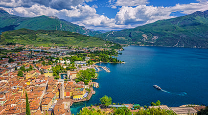 A scenic view of Lake Garda in northern Italy, surrounded by lush green hills and mountains. The foreground features the picturesque town of Riva del Garda, with its terracotta-roofed buildings nestled along the lake's edge. The deep blue waters of the lake reflect the vibrant sky, dotted with fluffy white clouds, as a boat glides across the serene surface.