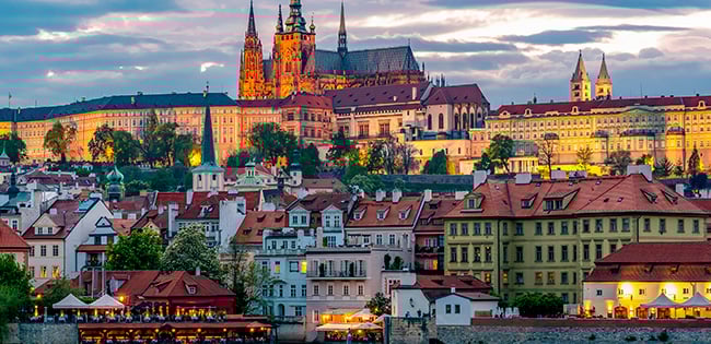 A scenic evening view of Prague, Czech Republic, featuring the illuminated Prague Castle atop a hill, with its gothic spires and historic architecture glowing warmly against a twilight sky. Below, colorful historic buildings with red-tiled roofs line the banks of the Vltava River, reflecting their lights in the calm water.