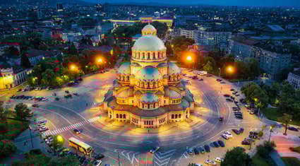 A stunning aerial view of Alexander Nevsky Cathedral in Sofia, Bulgaria, illuminated at night. The cathedral's golden domes and intricate architecture stand out against the darkened cityscape. Surrounding the cathedral are tree-lined streets and a circular plaza with parked cars, glowing under soft streetlights. The distant skyline of Sofia is visible under the deep blue evening sky.