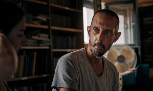 A person in a gray T-shirt sits in a dimly lit room, looking toward someone off-camera, surrounded by bookshelves.