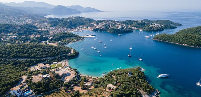 An aerial view of a serene coastal area in Greece, featuring a turquoise bay surrounded by lush green hills and small islands. Sailboats and yachts are anchored in the calm waters, while the shoreline is dotted with charming houses and buildings. The distant mountains add depth to the picturesque Mediterranean landscape under a clear blue sky.